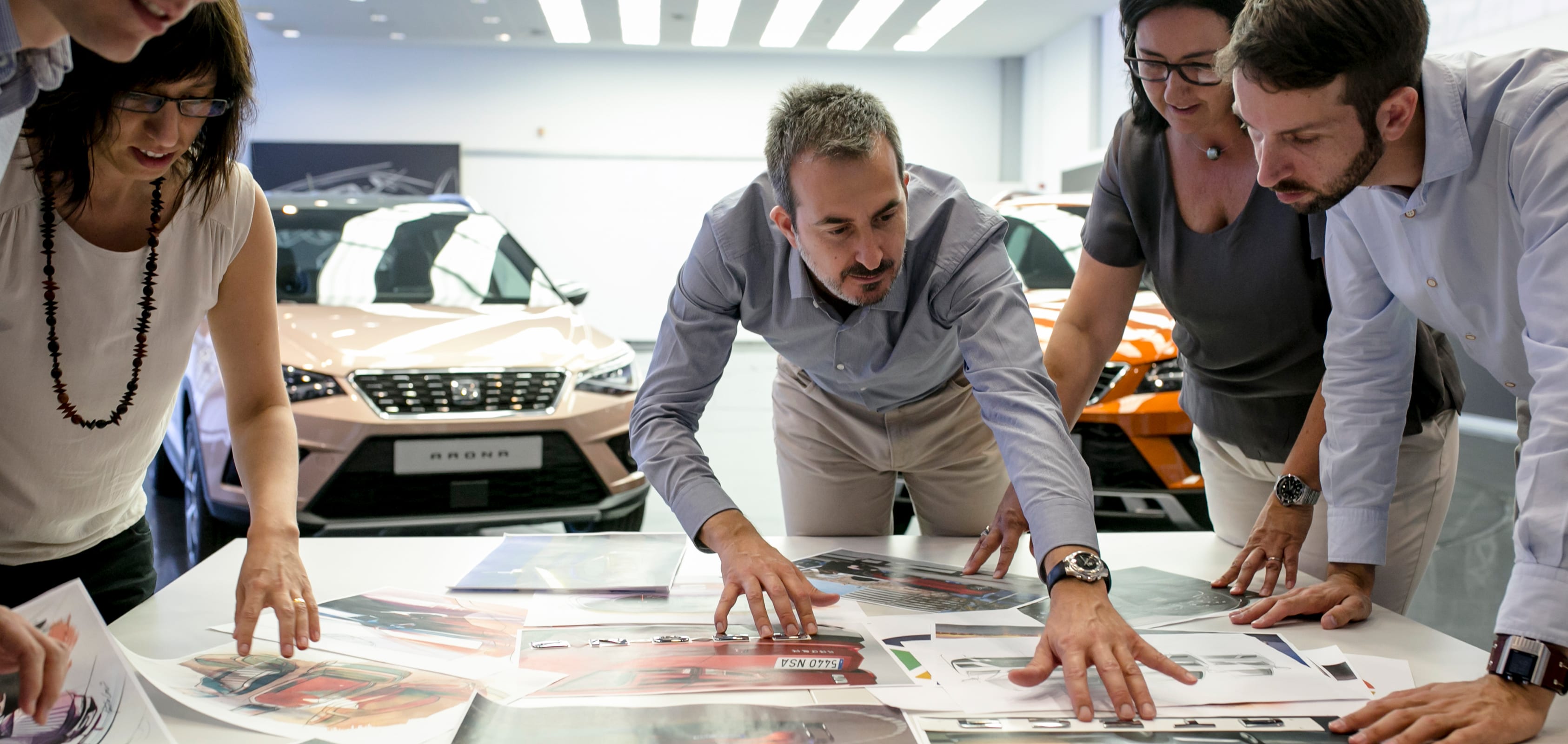 Four people inspecting photographs of SEAT car designs with SEAT Arona models in the background – SEAT Human Resources